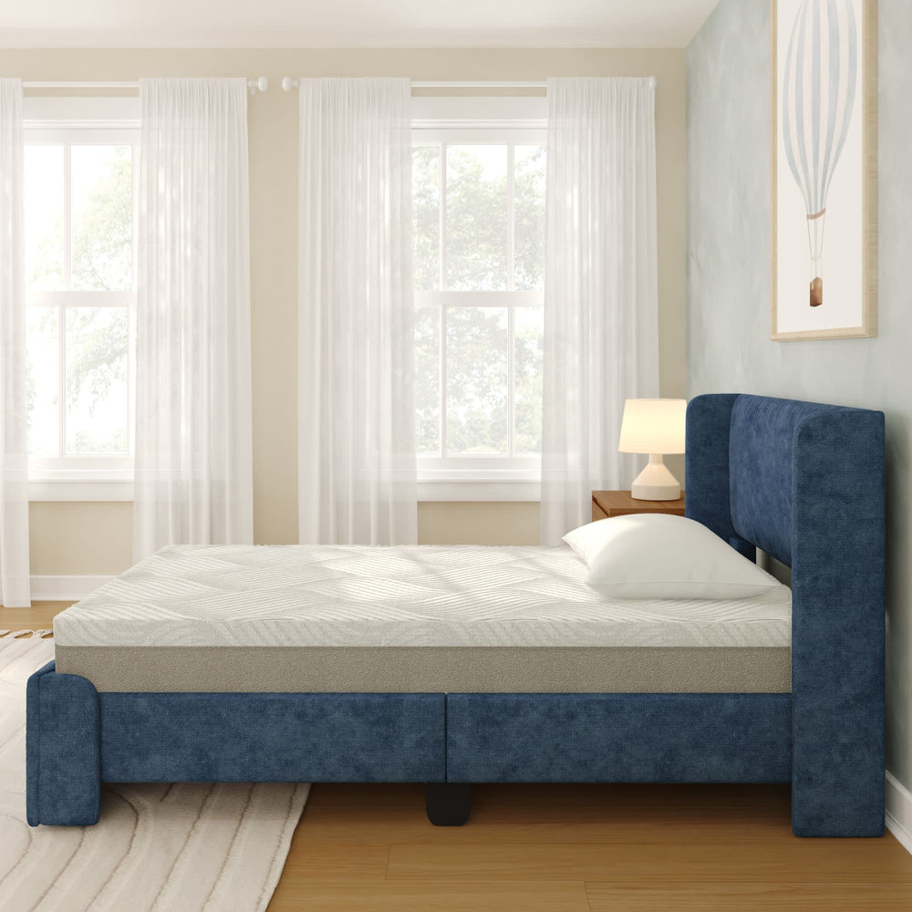 Blue upholstered bed with white mattress in a bright bedroom with sheer curtains.